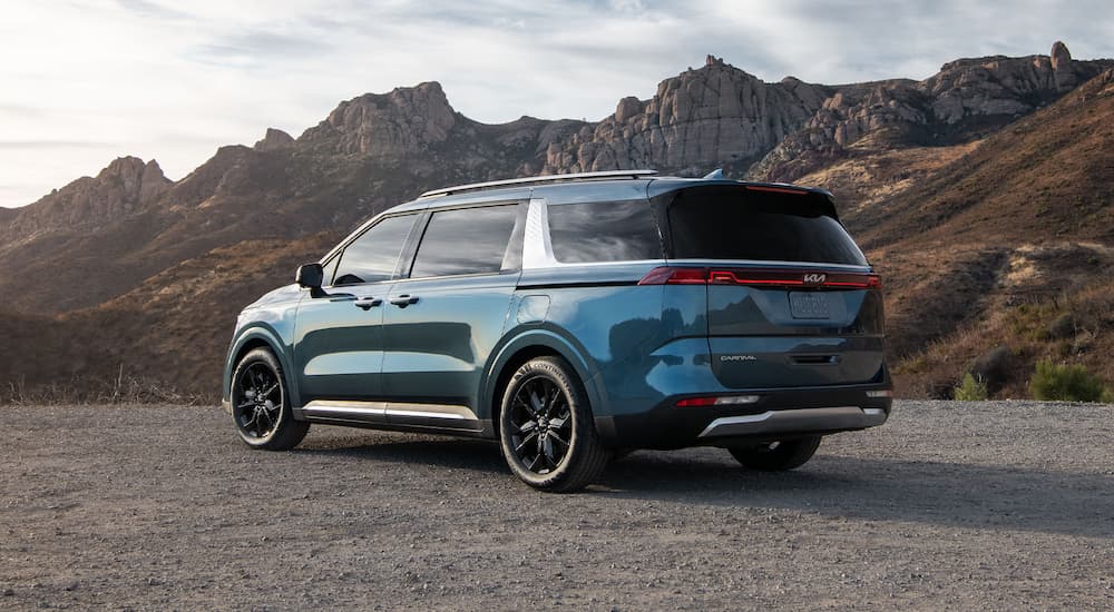 Rear view of a blue 2024 Kia Carnival parked near rocky mountains.