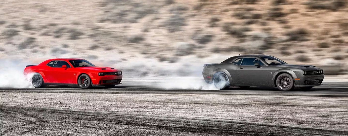 A red and a grey 2023 Dodge Challenger Hellcat are shown from the side while performing a burnout after leaving a Dodge dealer.