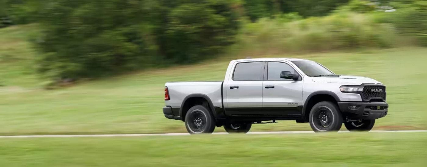 A silver 2025 Ram 1500 is shown from the side driving on an open road.